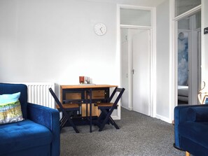 A room with a blue sofa, a wooden table with chairs, a white radiator, a clock, and a door leading to a staircase. - 2 bedroom accommodation in Woolacombe (Woolacombe)