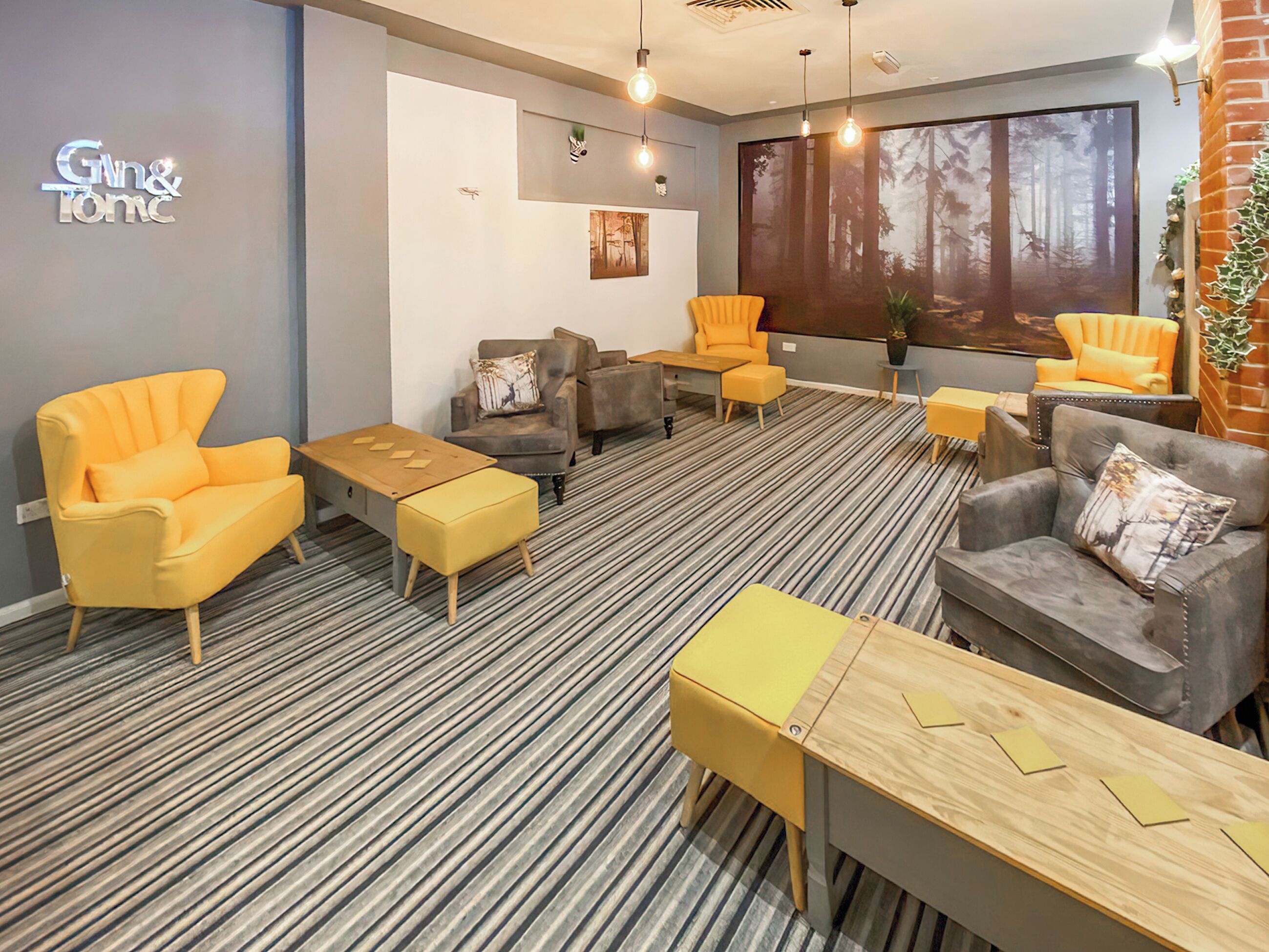 Ein moderner Lounge-Bereich mit Streifenboden, verschiedenen Sitzmöglichkeiten, darunter gelbe und graue Sessel, einem Holztisch und einem großen Fenster mit Blick auf einen Wald.