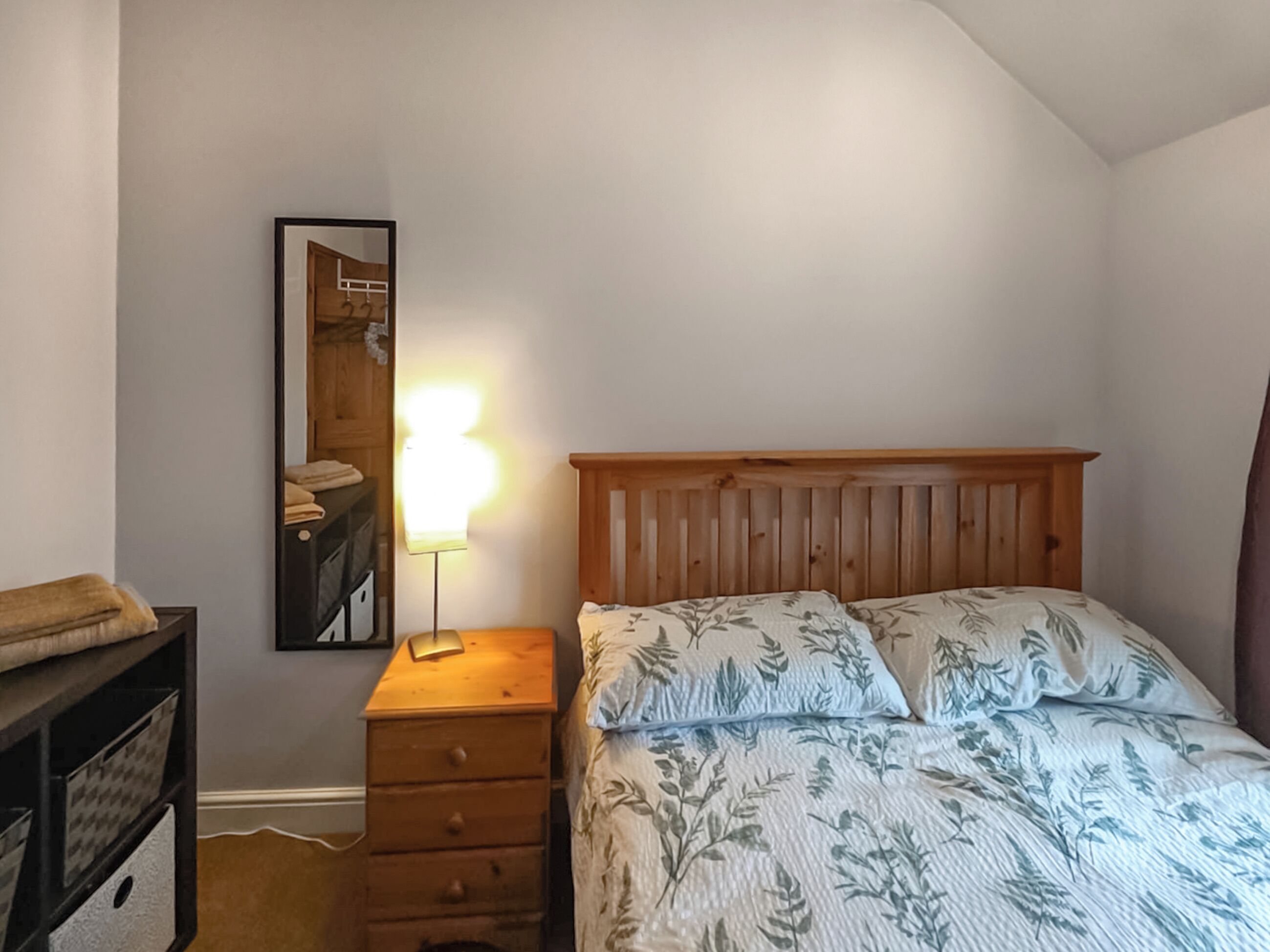 Un dormitorio con cabecera de madera, cama con ropa de cama floral, lámpara de noche, cómoda y un cuadro enmarcado en la pared.