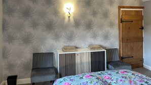 Una habitación con techo inclinado, cama con edredón floral, armario, dos sillas, una lámpara fijada a la pared y una puerta de madera.