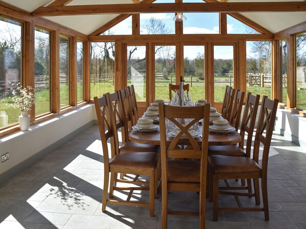A conservatory with a dining table set for six, surrounded by wooden chairs, large windows, and a view of the outdoors.