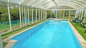 A covered swimming pool with a curved edge, surrounded by a glass enclosure, and a grassy area with lounge chairs.