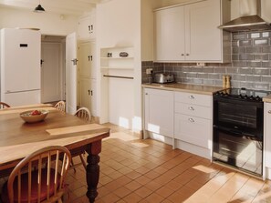 A kitchen with white cabinets, a wooden dining table, chairs, a stove, and a tiled backsplash. - 6 bedroom accommodation in Trebetherick, near Wadebridge (Trebetherick, near Wadebridge)