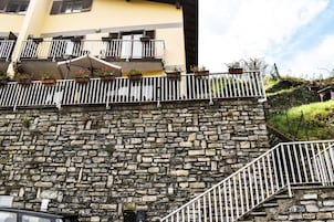 Un muro de piedra con barandilla metálica, un edificio amarillo con balcones y una escalera.