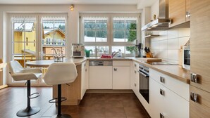 A modern kitchen with white cabinets, a dishwasher, and a stove. There are two bar stools and a dining table.