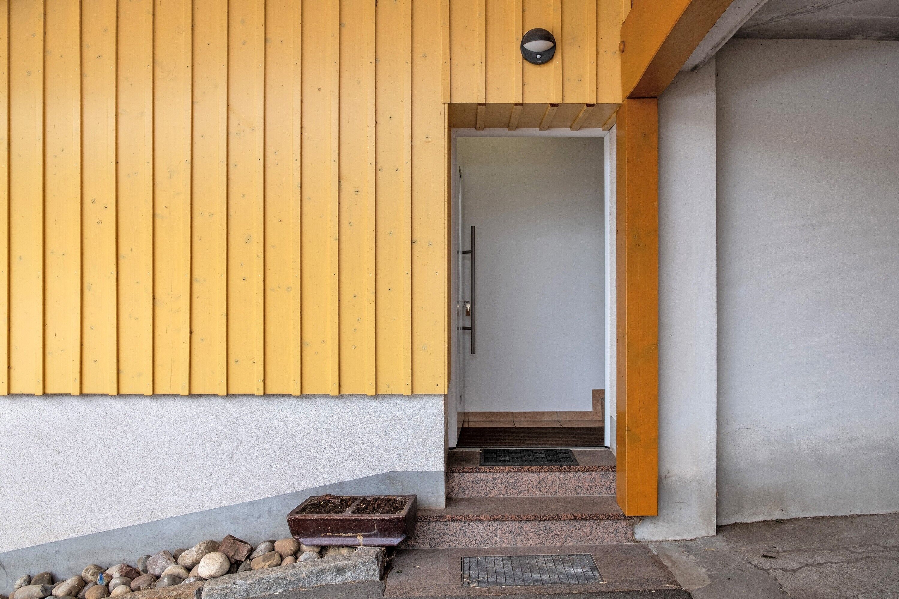 A wooden exterior wall with a door, steps leading to a threshold, and a small outdoor area with rocks and a planter.