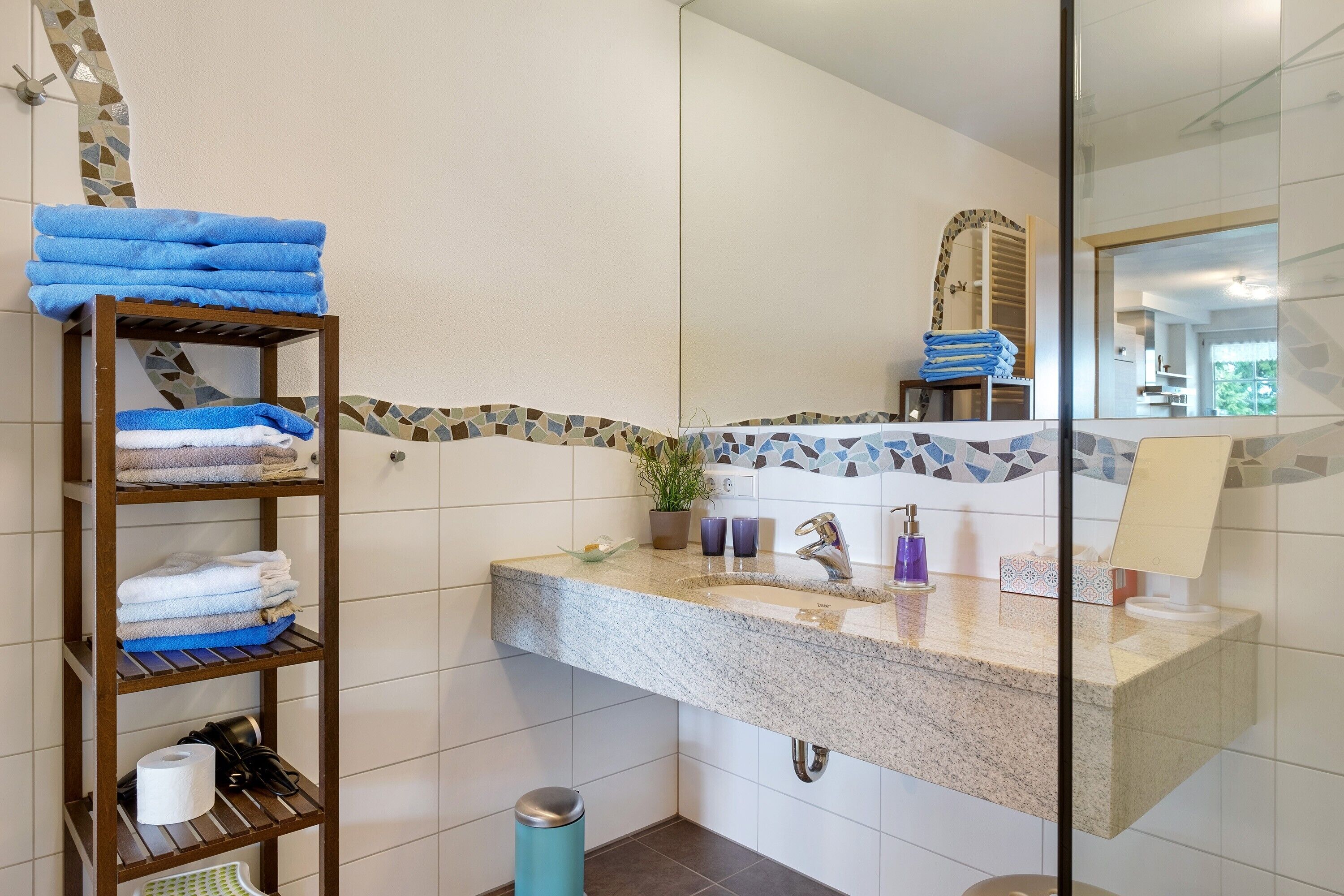 Un baño con encimera de granito, un lavamanos con grifo, un espejo, un estante con toallas dobladas y un cubo de basura.