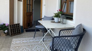 Un pequeño patio con una mesa blanca y dos sillas, una planta en maceta y una ventana con persianas.