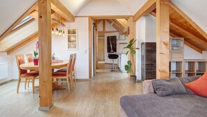 Een ruime loft met een hoge plafond, houten balken, een eethoek met stoelen en een tafel, een keuken met kasten, en een gezellige zithoek met kussens.