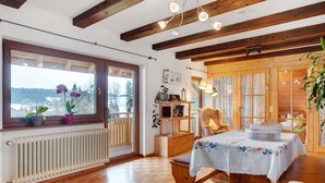 Une pièce avec un plancher en bois, un radiateur, une table recouverte d’une nappe à motifs floraux et une fenêtre donnant sur des arbres et des montagnes.