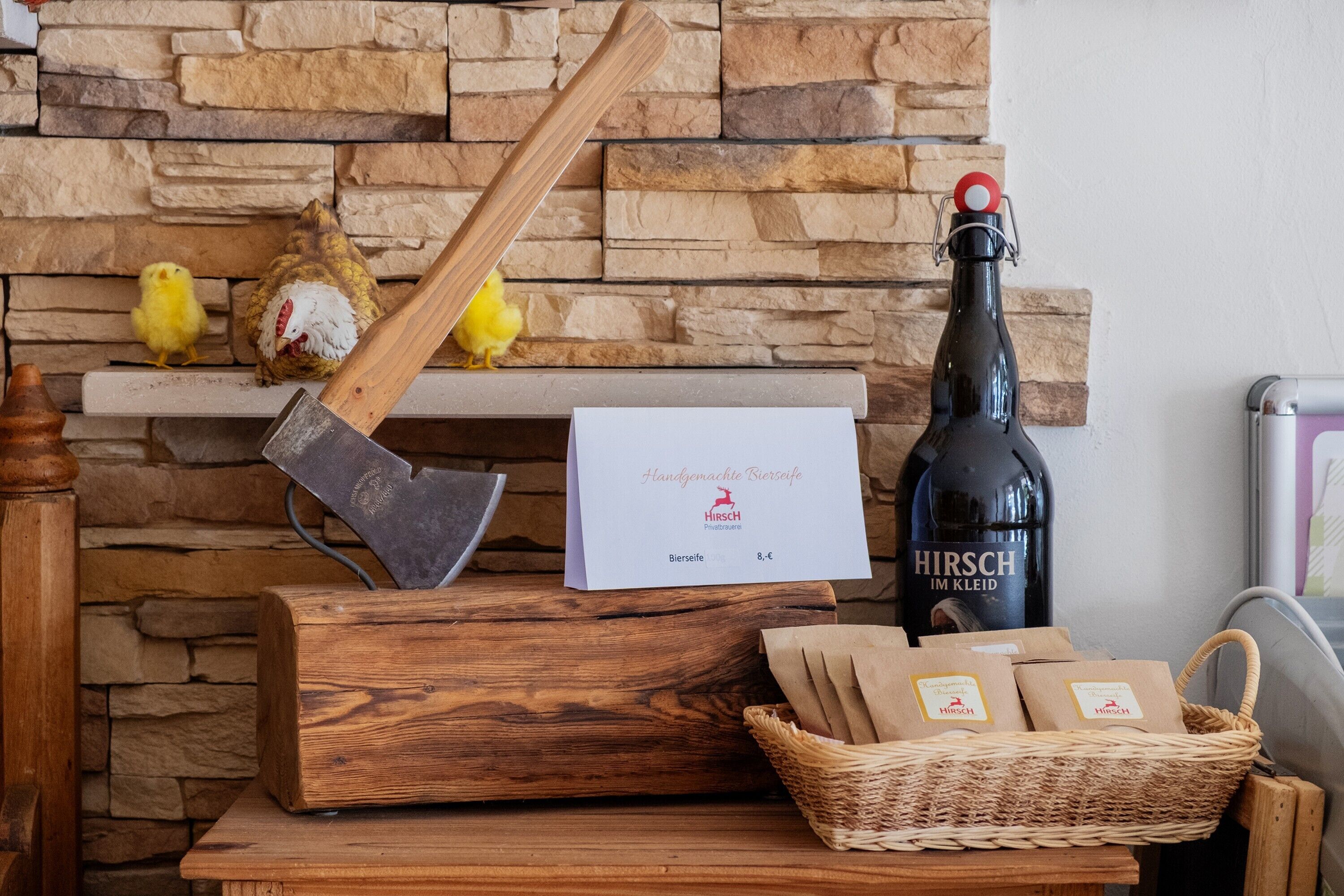 Un muro de piedra, un tronco de madera para cortar, una botella de cerveza Hirsch y un cartel con precios.