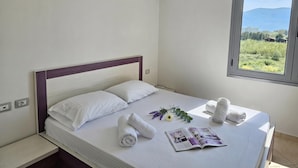 A neatly made bed with white linens, a window with a view of greenery and mountains, and a wall-mounted air conditioner. - Apartment With Pool View 001 (Orikum)