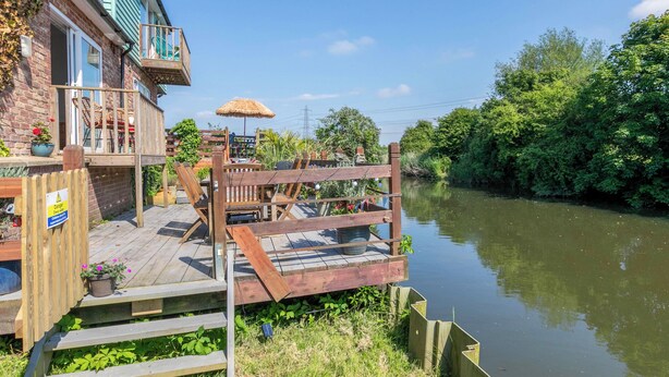 Eine Holzterrasse mit Gartenmöbeln, einem Reetdachschirm und Blick auf einen Fluss.