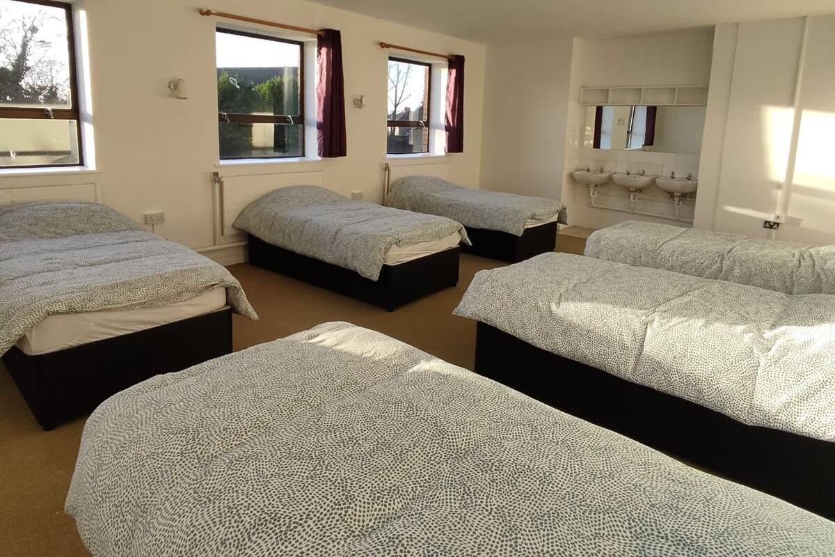 A room with four beds, a bathroom with two sinks, and windows with curtains.