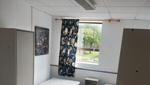 Ein Schlafraum mit zwei Einzelbetten, einem kleinen Tisch, einem Kleiderschrank und einem Fenster mit Blick auf Grünflächen.