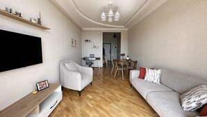 A modern living room with a flat-screen TV, a grey sofa, a wooden coffee table, and a dining area with a table and chairs. - Playstation 5 + Panoramic City view Apartment (Baku)