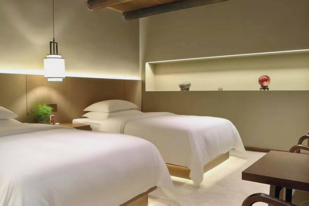 A hotel room with two beds, a bedside table, a small desk, a lamp, and a shelf with decorative items.