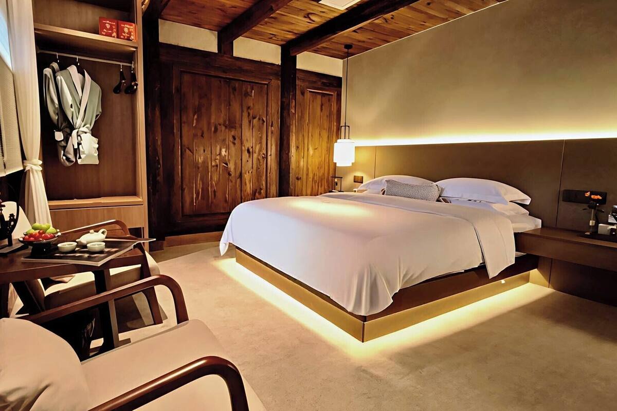 A hotel room with a wooden ceiling, a bed with white bedding, a bedside table with a lamp, a chair, and a small table with a bowl of fruit.