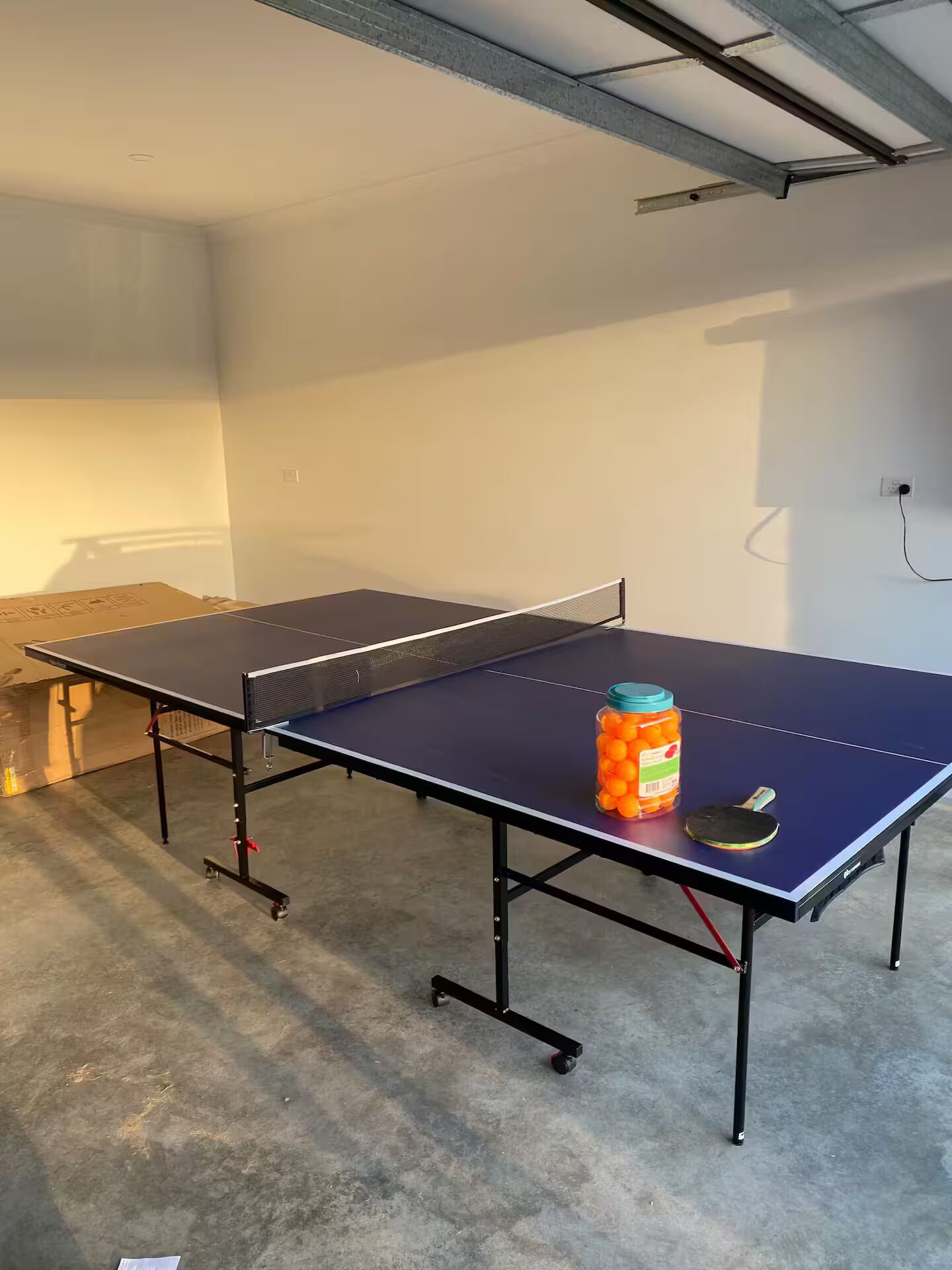 A ping pong table with a jar of oranges and a paddle in a room with a concrete floor and a white ceiling.