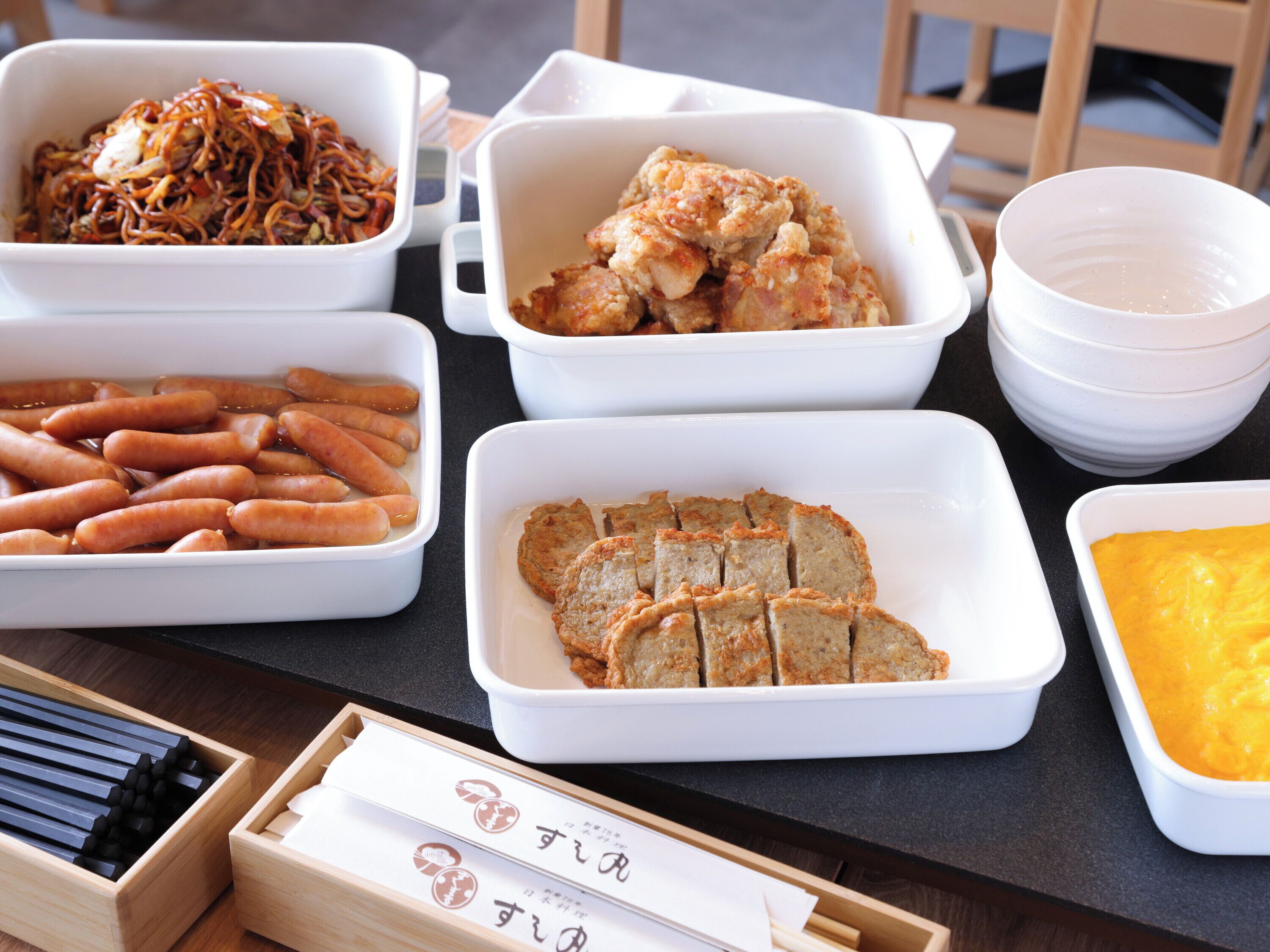 A table with various food items in white containers, including sliced bread, sausages, and a dish with a yellow sauce.