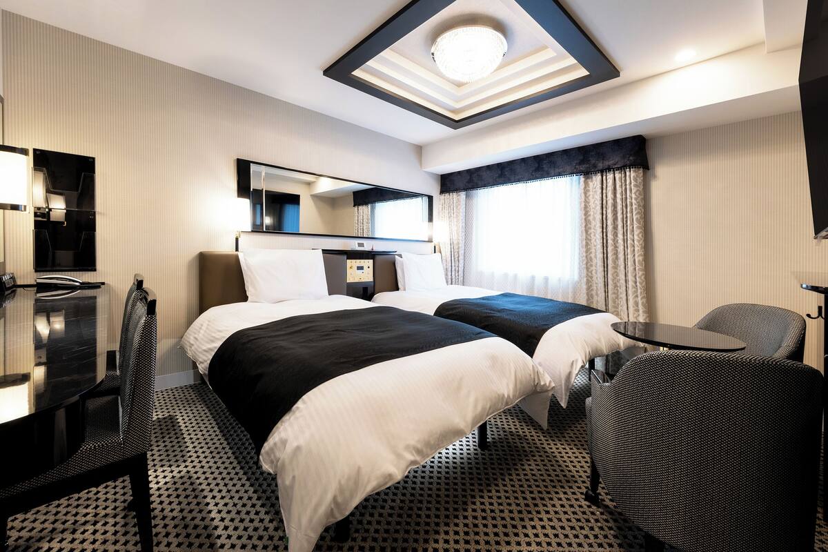 A hotel room with two beds, a desk, a chair, a mirror, and a ceiling light fixture.