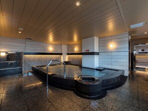 Un área de piscina interior moderna con un jacuzzi central, zona de duchas y un reloj digital que muestra la hora.