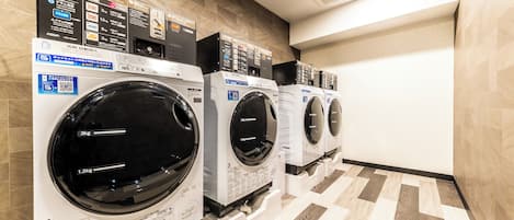 A row of front-loading washing machines in a hotel room with a tiled floor and a plain wall.