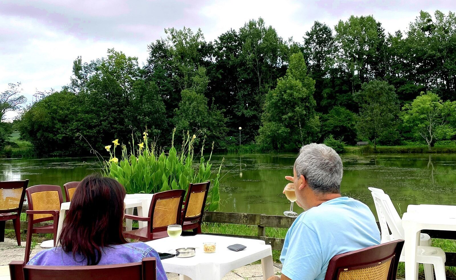 A couple dining at an outdoor table with chairs, overlooking a pond surrounded by trees.