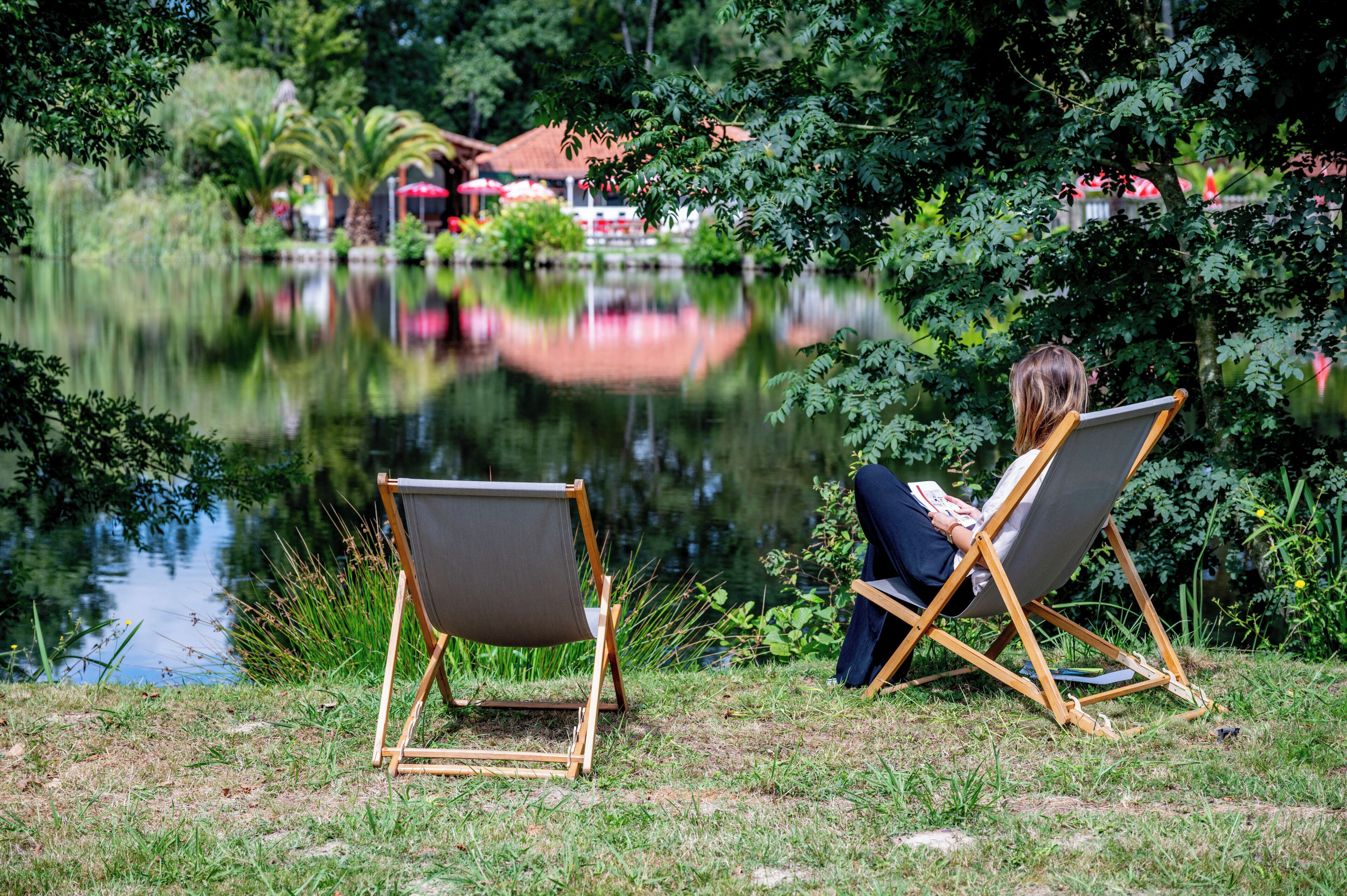 A person sitting on a foldable chair by a calm lake, surrounded by lush greenery and a distant building with a red roof.