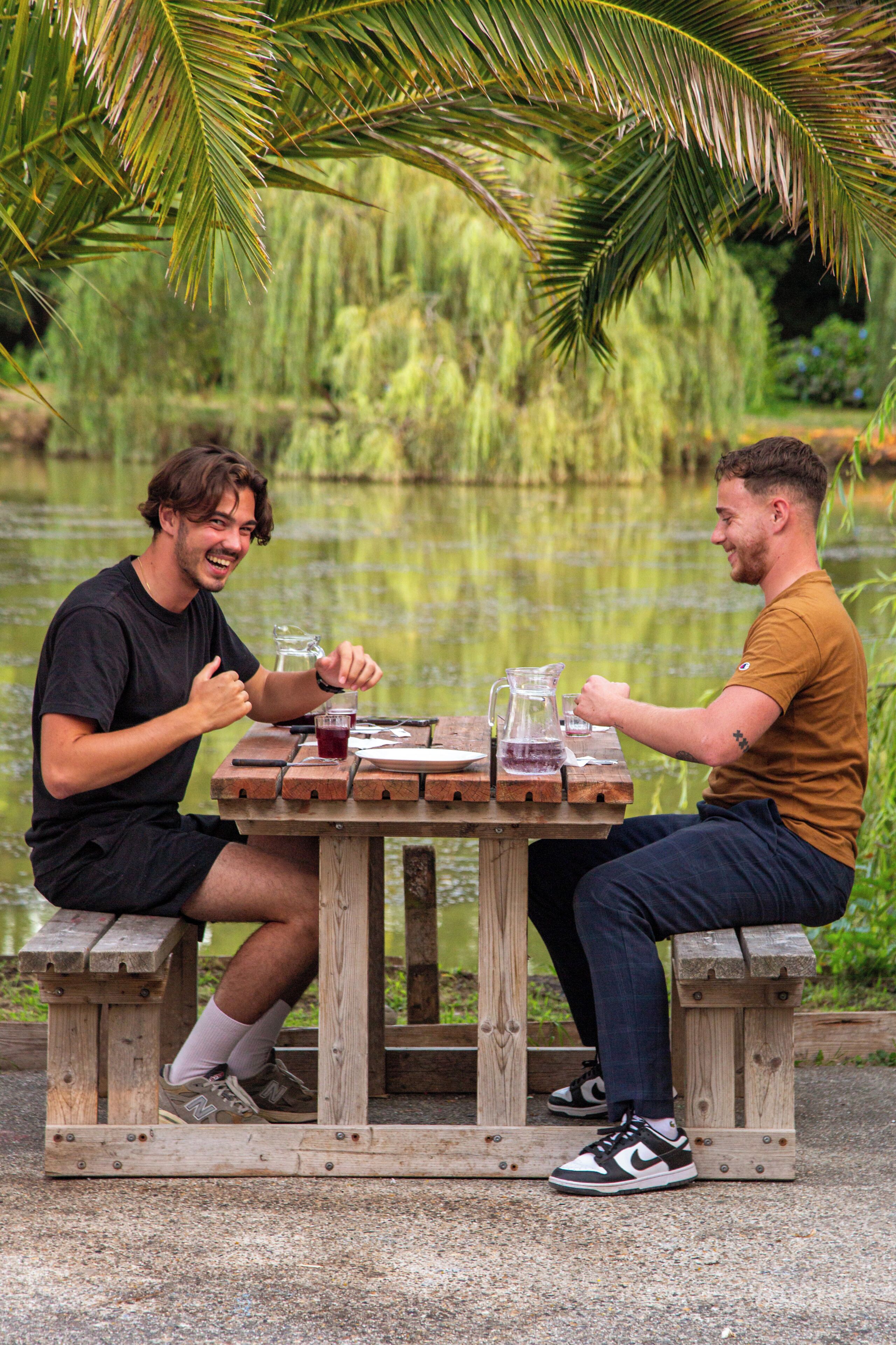 Deux personnes sont assises à une table en bois, devant une carafe et des verres, à l’extérieur, près d’une étendue d’eau.