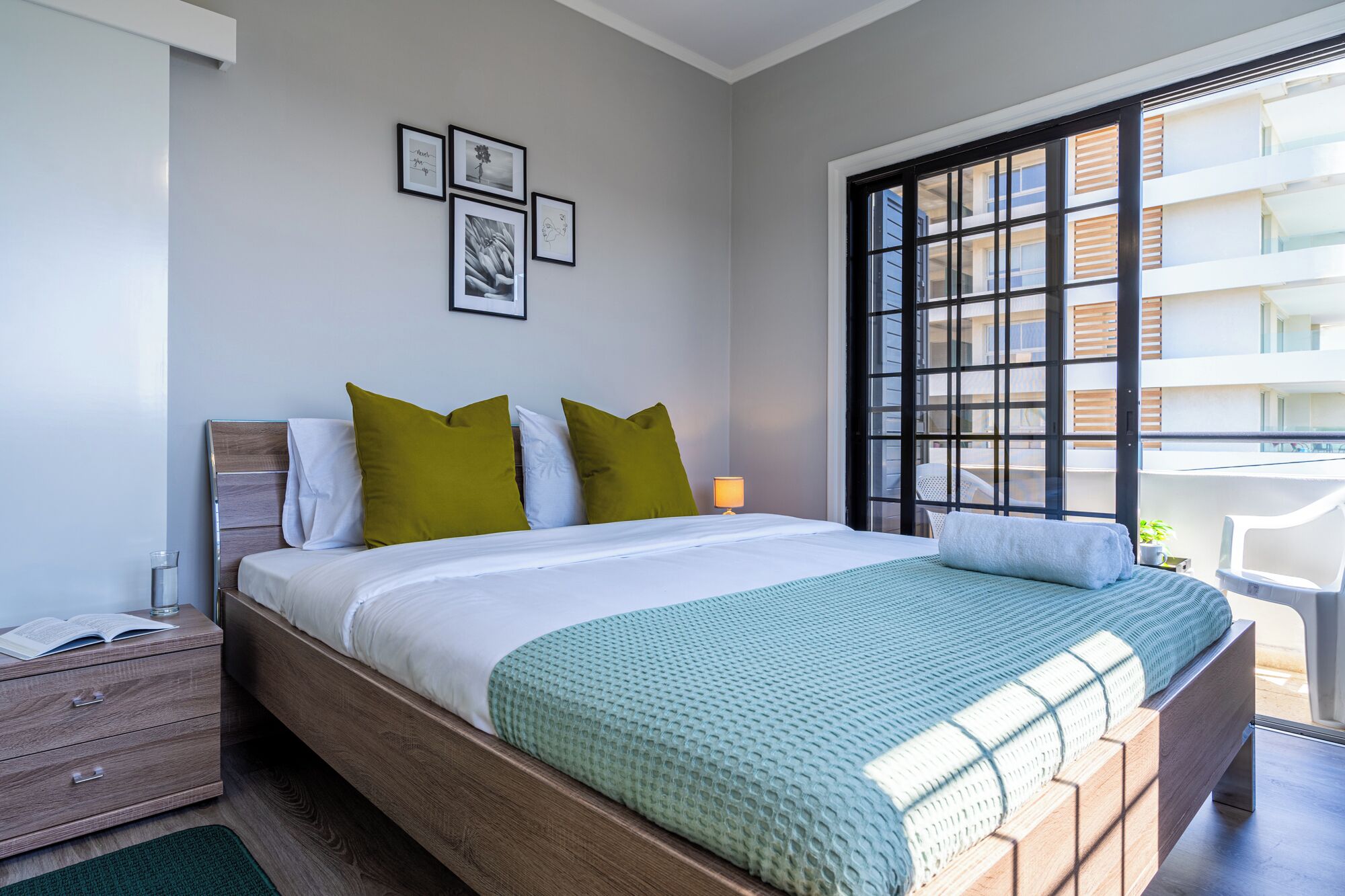 Ein modernes Schlafzimmer mit einem Holzbett, grünen Kissen, einem Nachttisch mit Lampe und Blick auf die Stadt durch große Fenster.