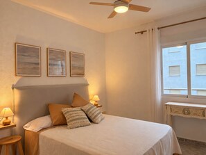 A bedroom with a bed, a ceiling fan, a window with curtains, and wall-mounted picture frames. - Ninomad 02 Beachfront - Near Gandía (Daimús)