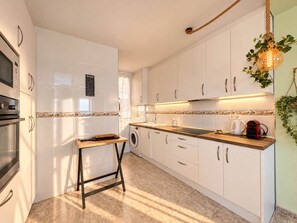 A modern kitchen with white cabinets, a wooden countertop, a washing machine, and a hanging light fixture. - Ninomad 02 Beachfront - Near Gandía (Daimús)