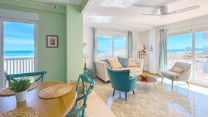 Een ruime woonkamer met uitzicht op het strand, een eettafel met een plant en een plafondventilator.