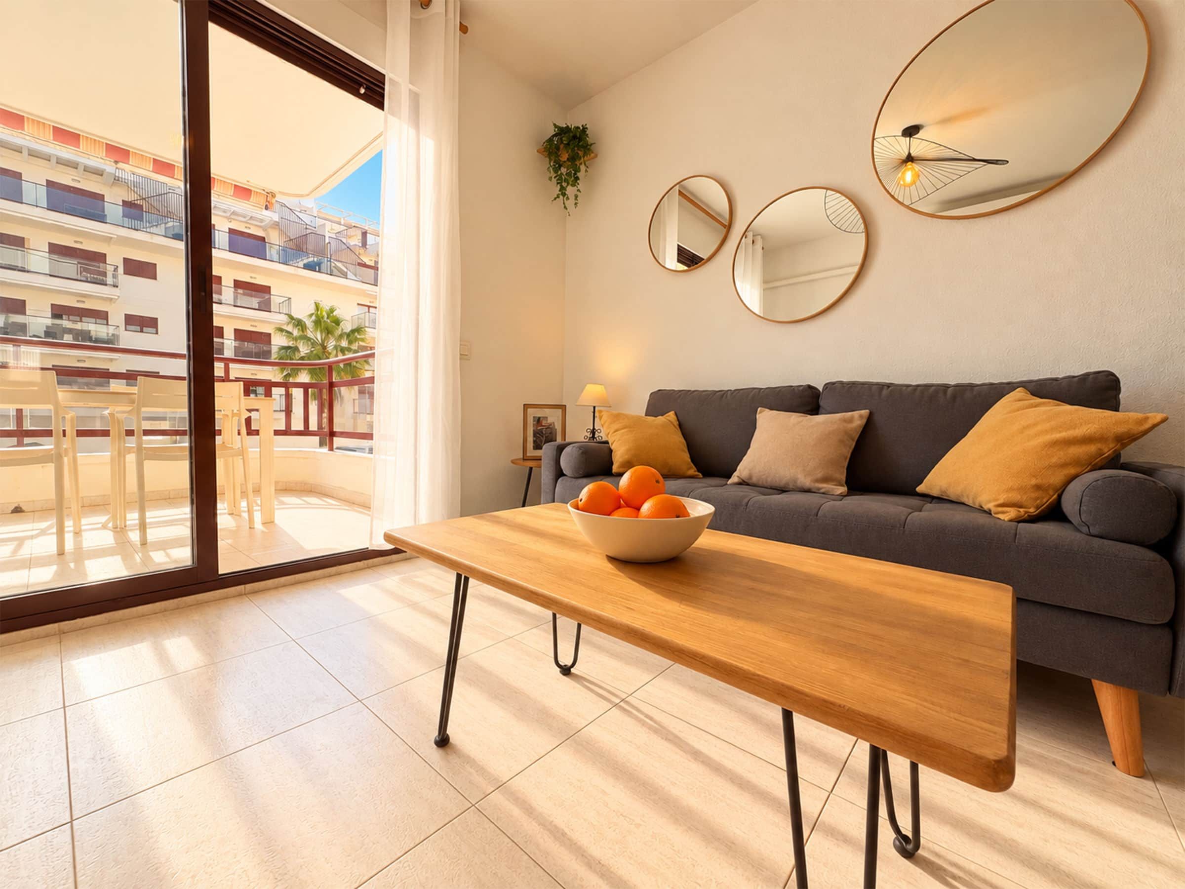 Un salon moderne avec un canapé, une table en bois et un saladier rempli d’oranges. Deux miroirs sont accrochés au mur, et un balcon offre une vue sur un immeuble.