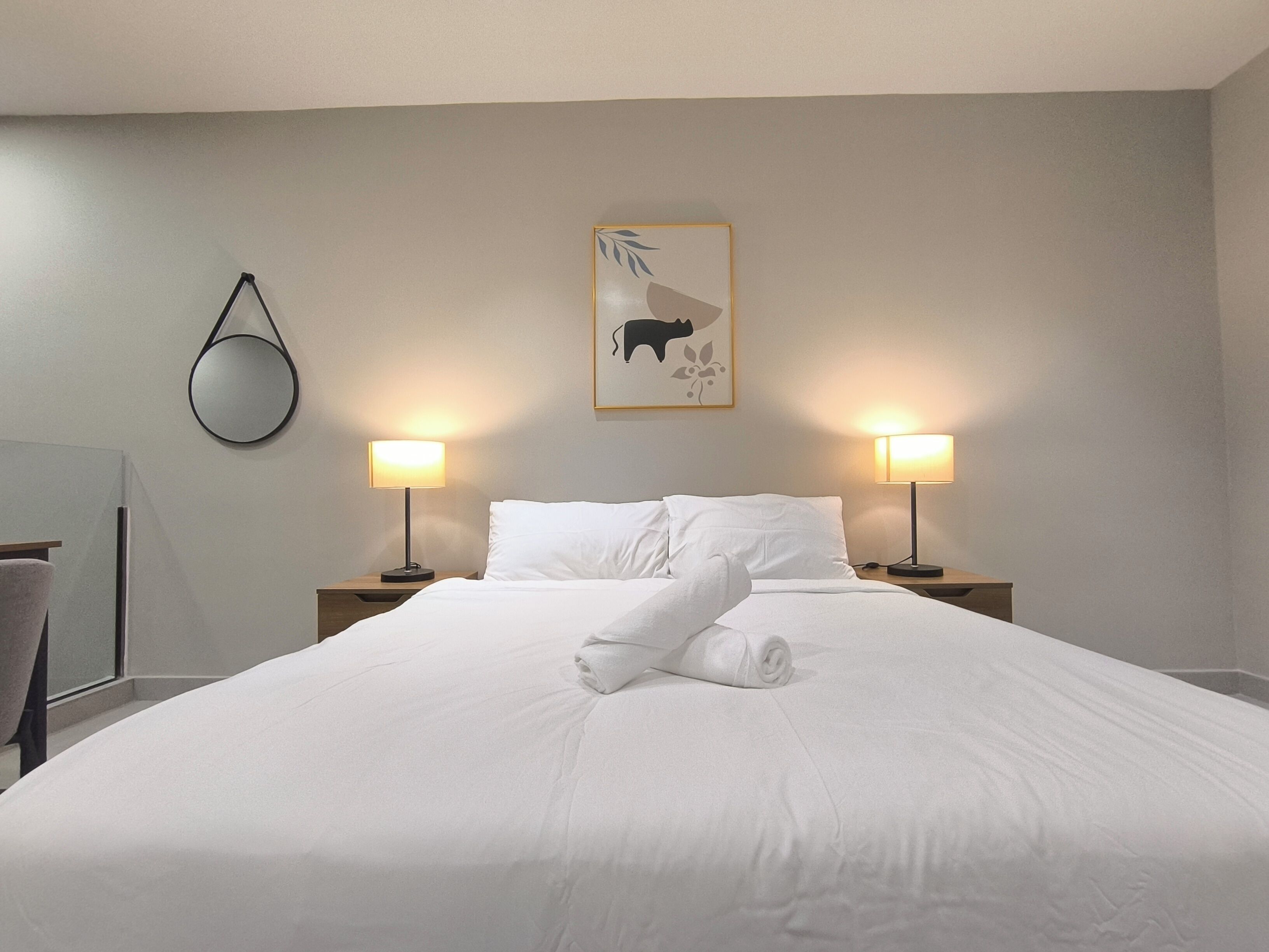 A neatly made bed with white linens, two bedside lamps, a wall-mounted mirror, and a framed picture above the bed.