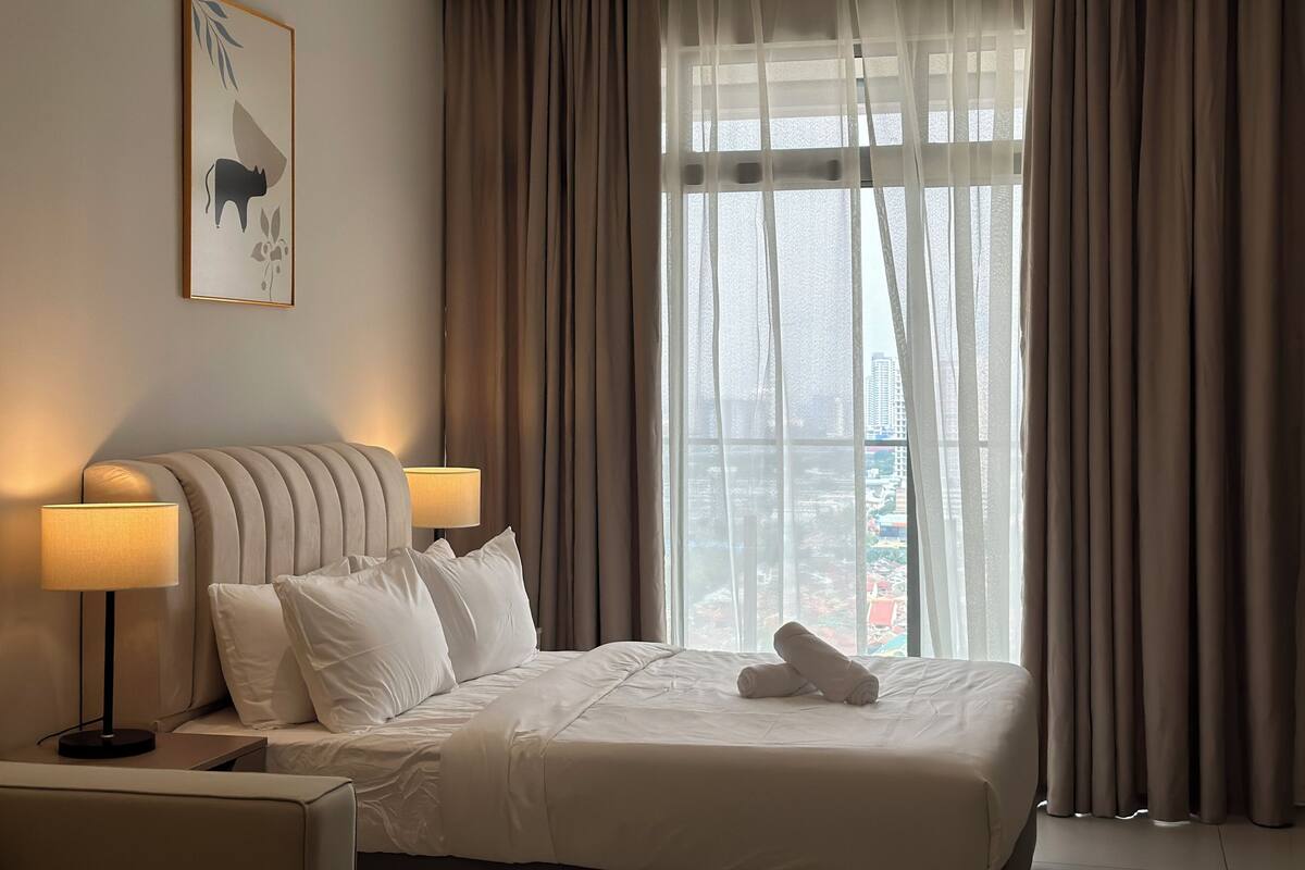 A hotel room with a bed, two bedside lamps, a window with curtains, and a cityscape view.