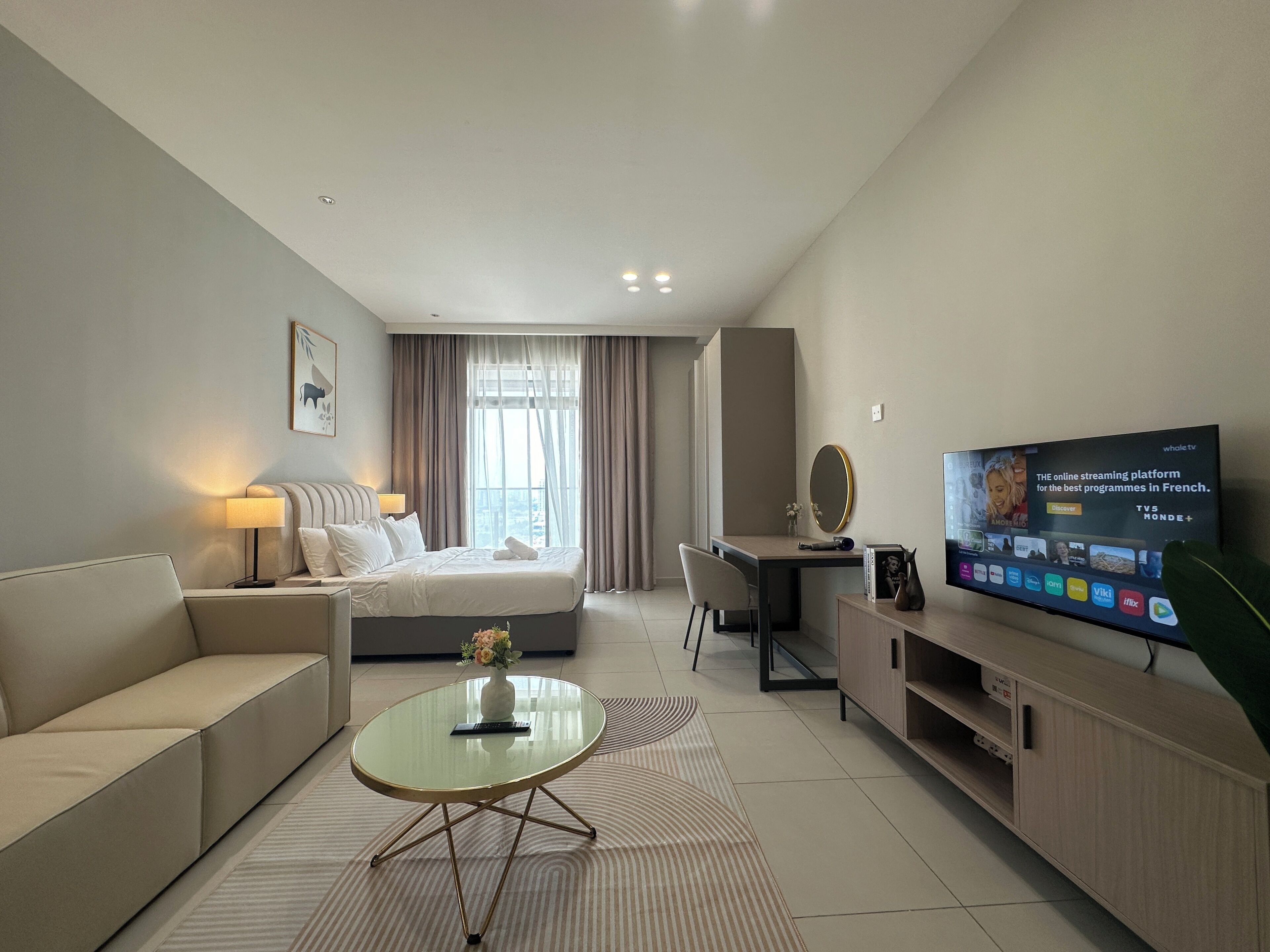 A modern hotel room with a bed, sofa, desk, TV, and a view of the city.