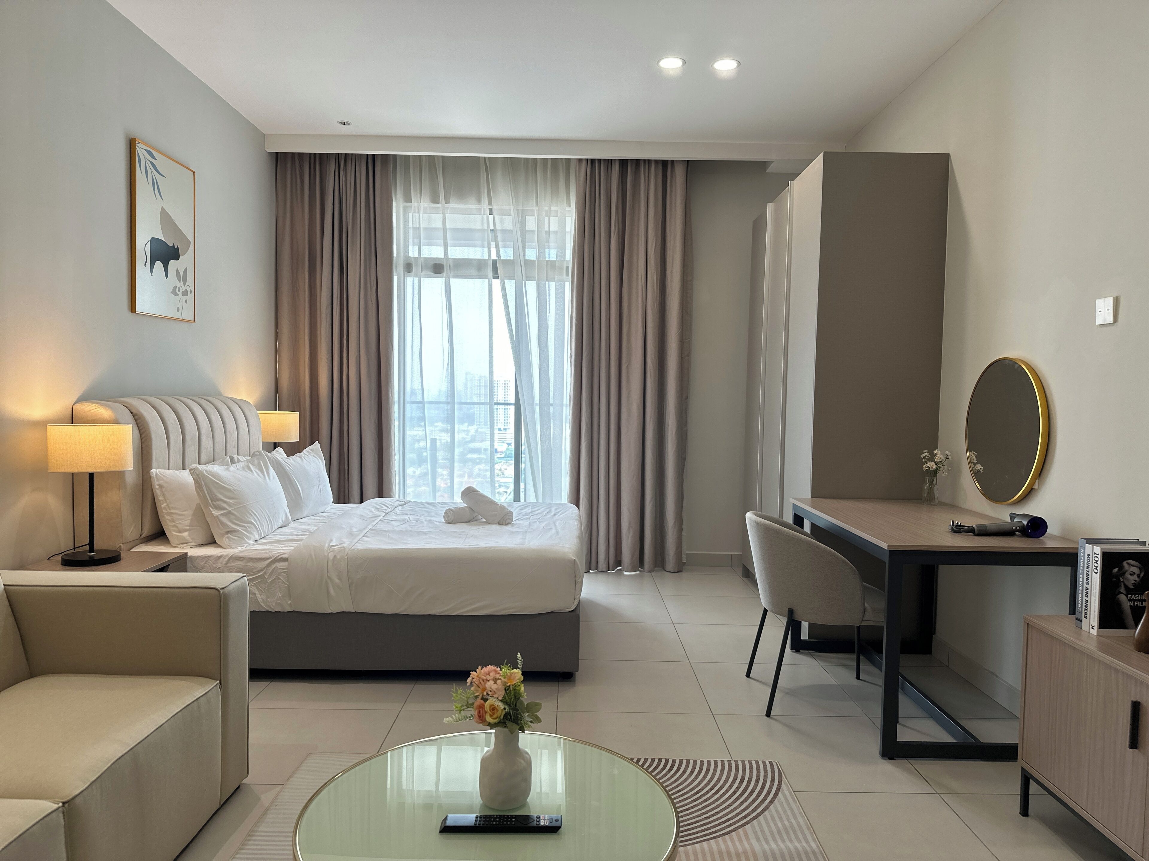 A hotel room with a bed, bedside lamps, a desk, a chair, a round glass coffee table, and a vase with flowers.