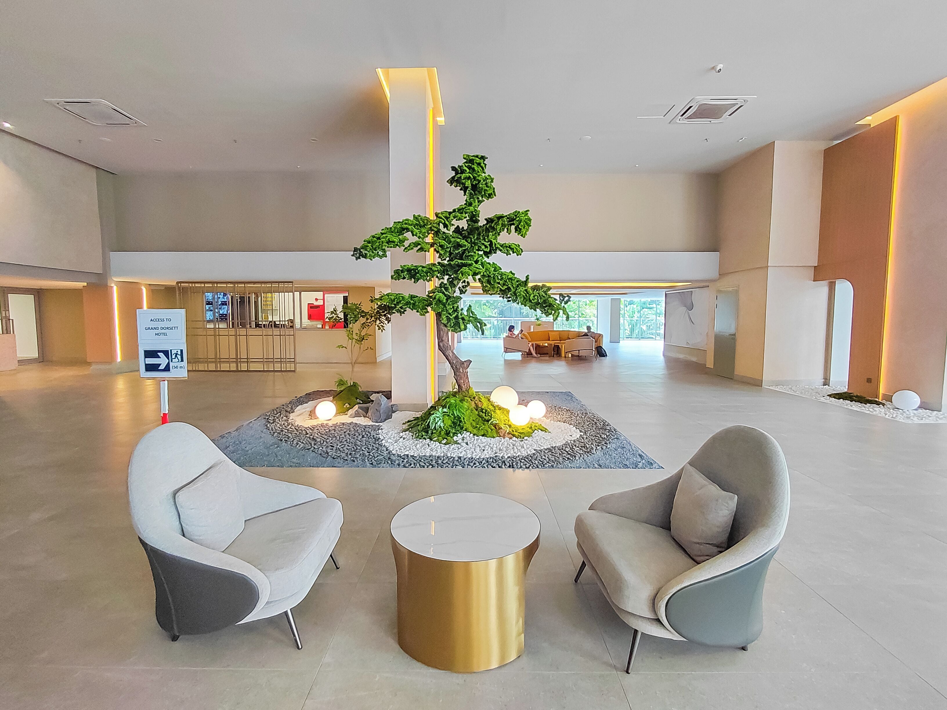 A lobby with a reception desk, two grey armchairs, a small round table, and a decorative tree centerpiece.