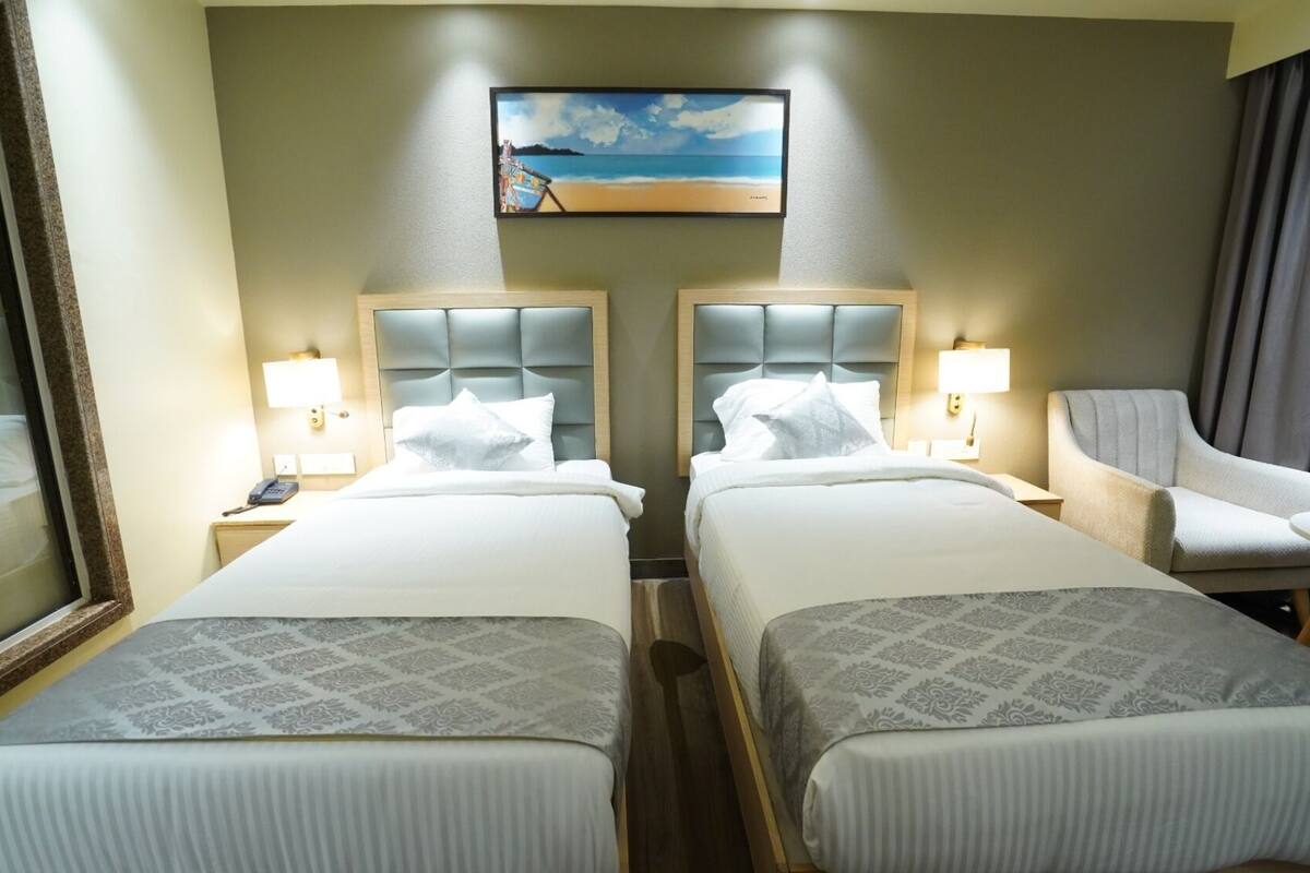A hotel room with two beds, a painting of a beach scene, a chair, and a nightstand with a lamp.