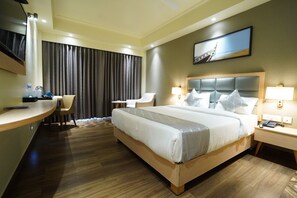 A hotel room with a bed, a desk, a chair, a TV, and a painting on the wall. - Zenvora Heights (A Unit of Aarokan Heights) (Puri)