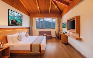 Quarto de hotel com cama, sofá, escrivaninha, televisão e uma janela grande com vista para as montanhas.