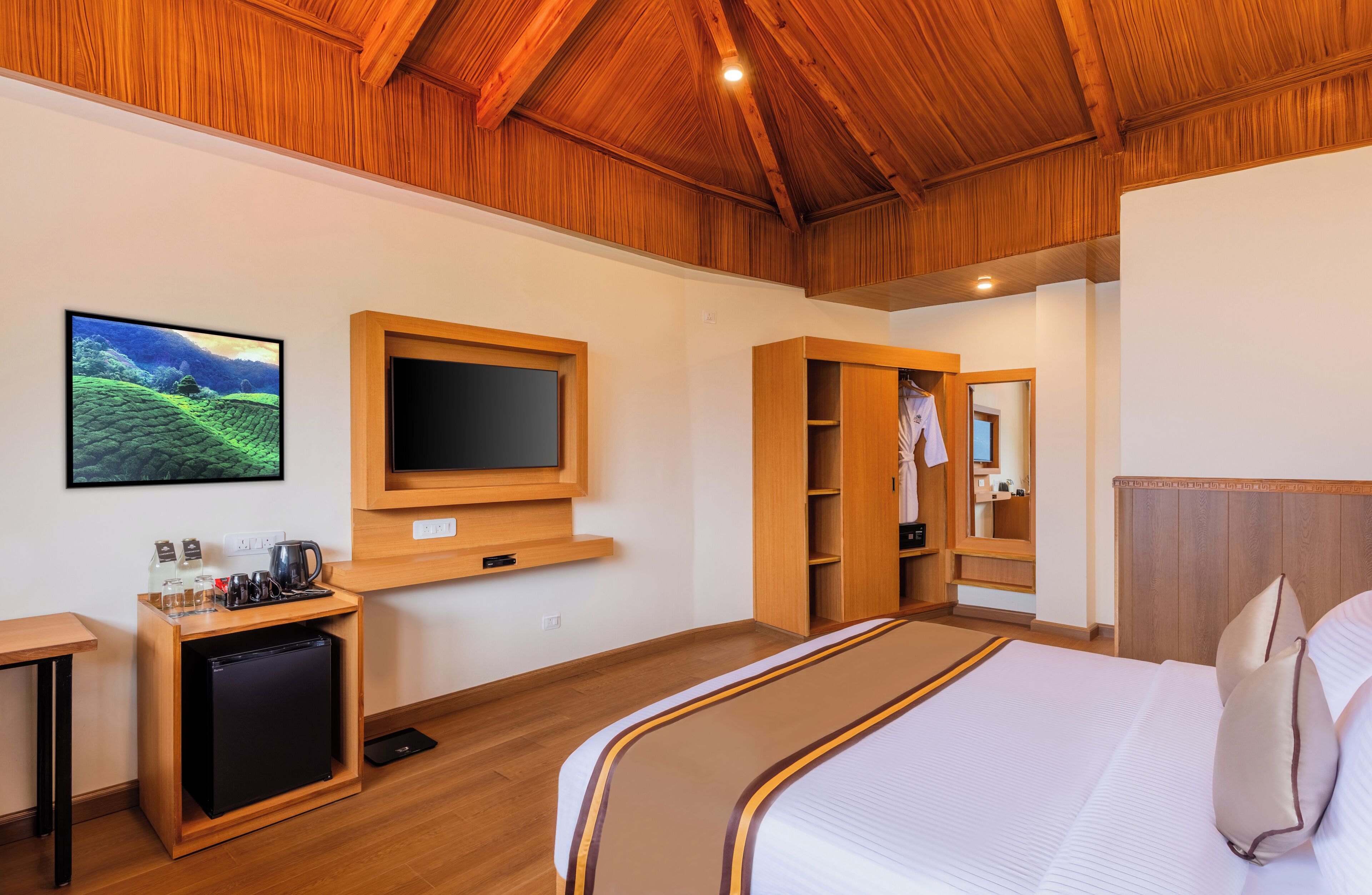 A hotel room with a bed, a television mounted on the wall, a small kitchenette, and a wooden ceiling.