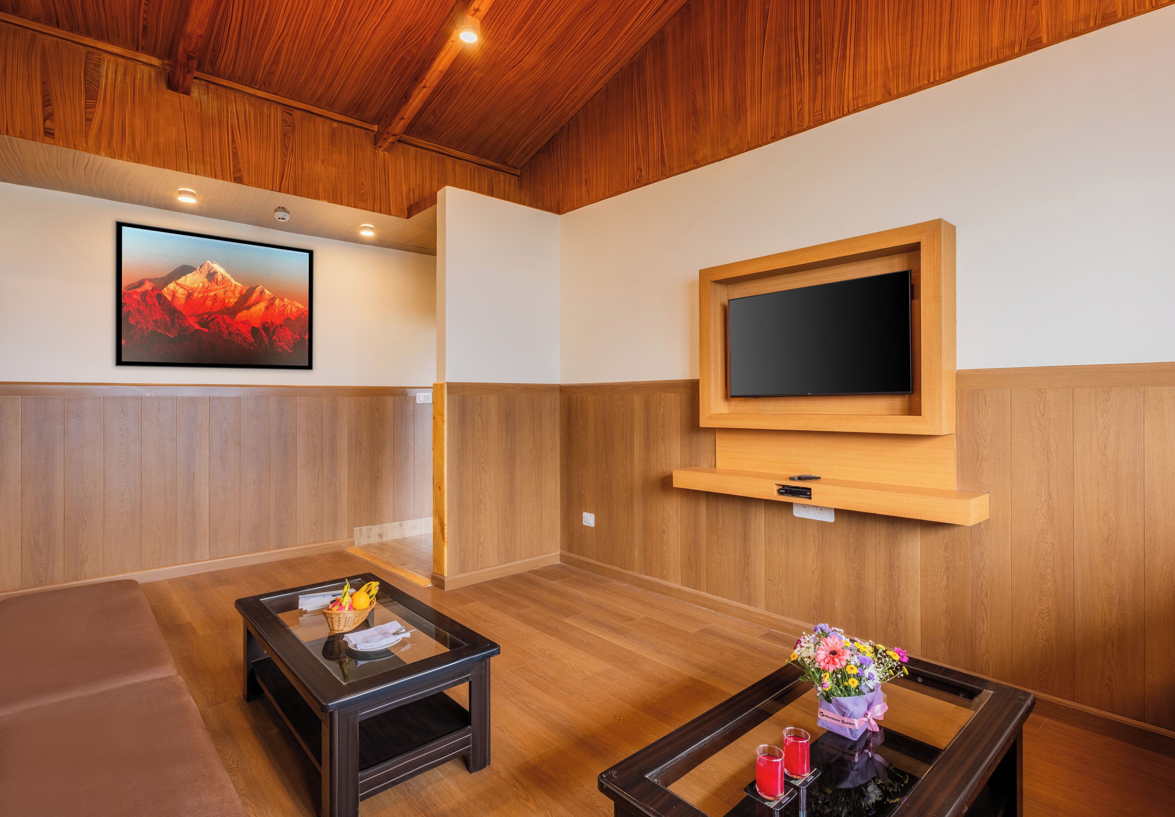 A room with a wooden ceiling, a mounted TV, a framed picture, a sofa, and a coffee table with a vase of flowers.
