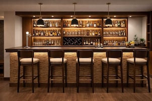 Uma área de bar com balcão de madeira, várias prateleiras de garrafas e banquetas.