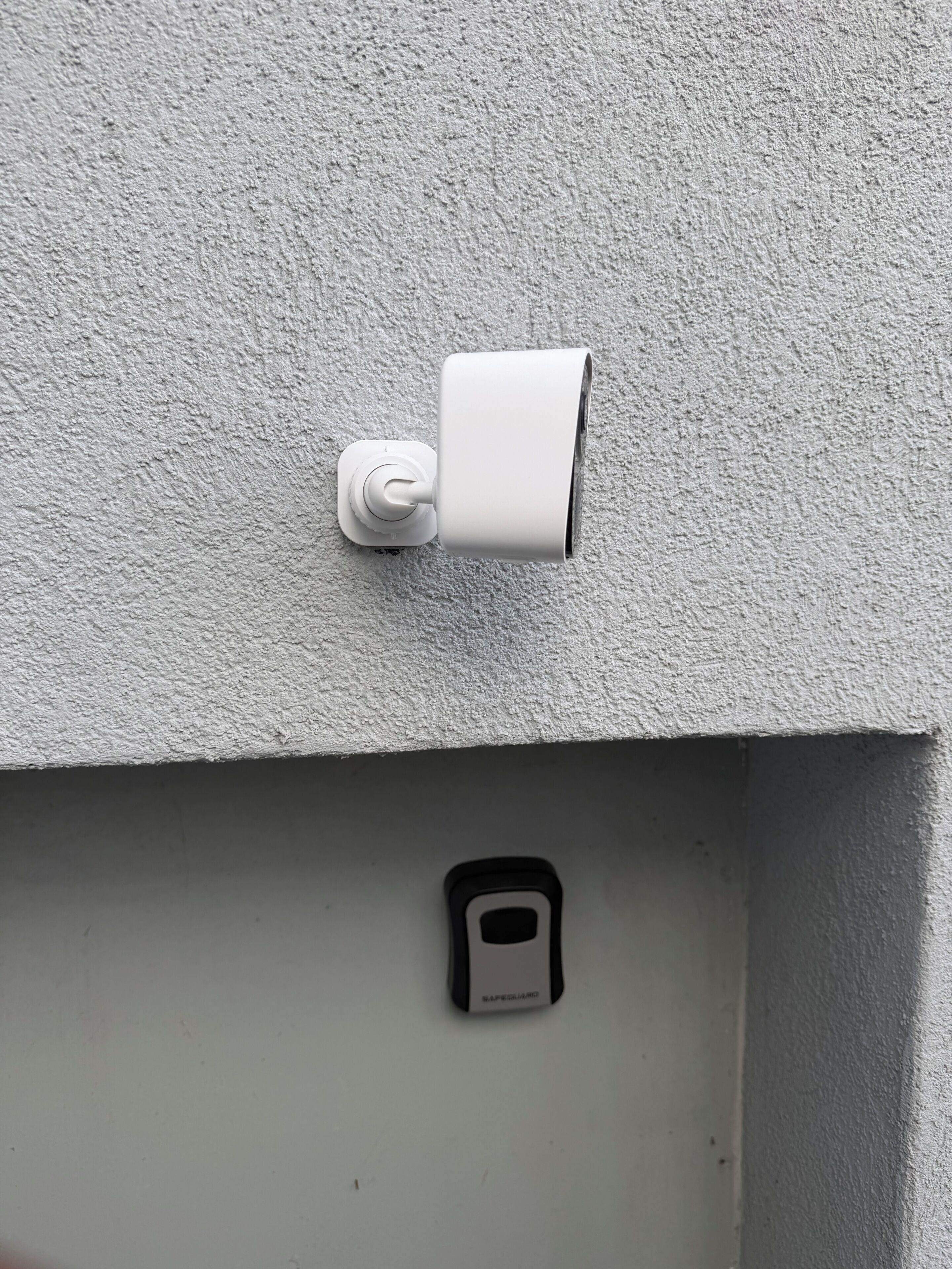 A security camera mounted on a textured wall, a smoke detector below it, and a small, rectangular object on the wall.