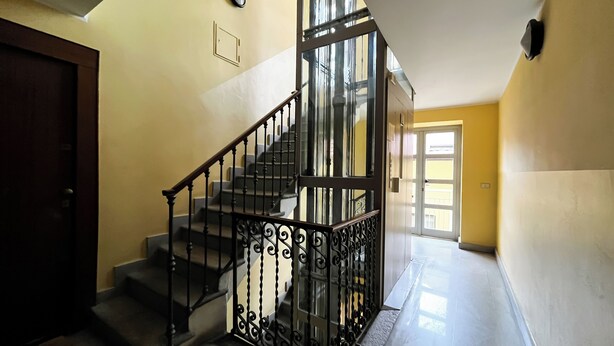 A staircase with ornate black railings, a door on the left, and a bright hallway leading to an open door on the right. - Sirtori Milano (Milan)