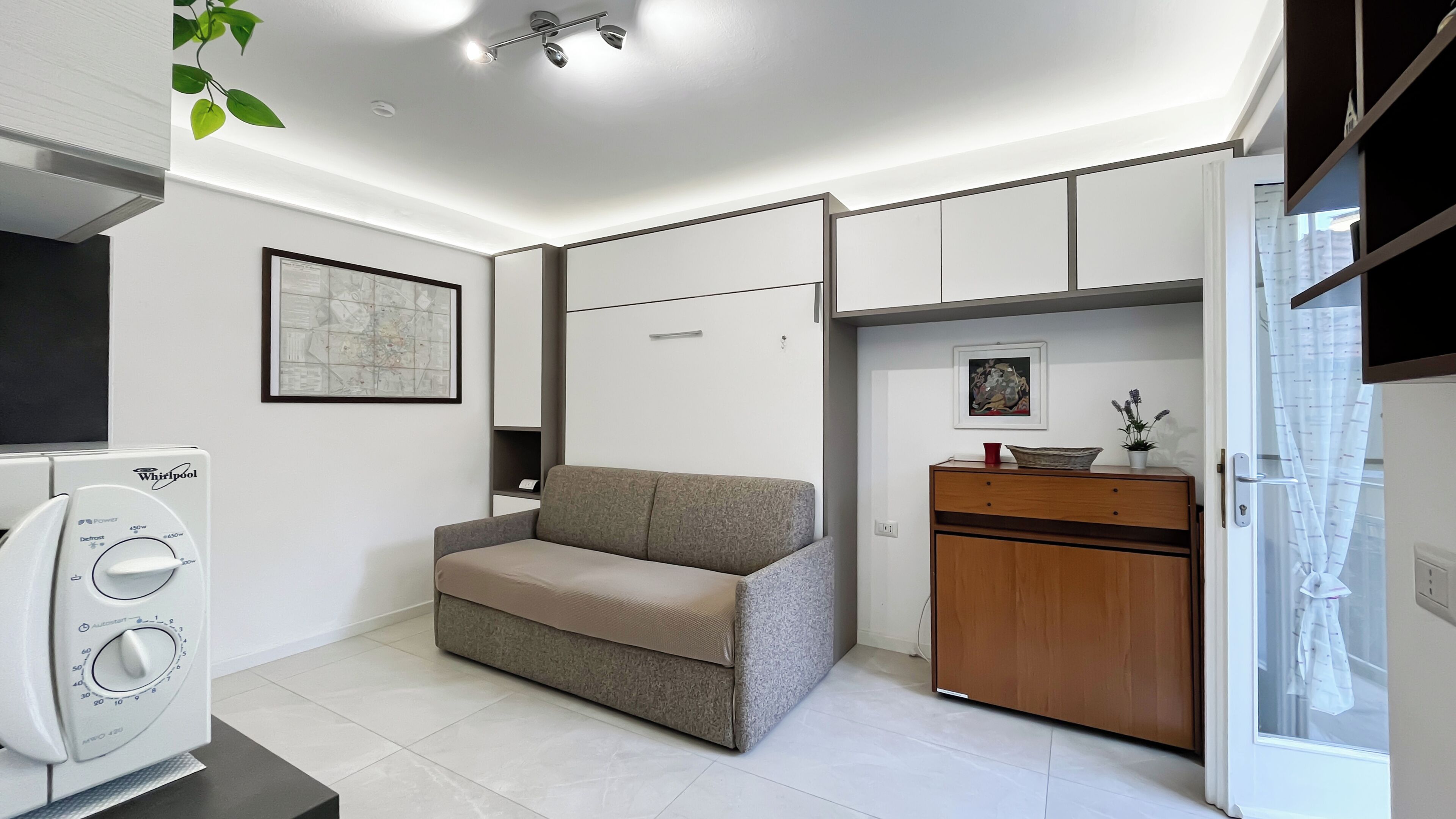 A compact room with a sofa, a wooden cabinet, a small table with a plant, and a Whirlpool appliance.