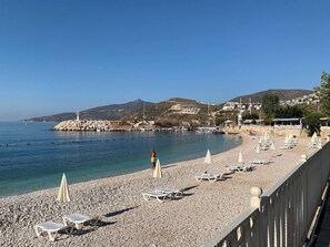 A pebble beach with lounge chairs and umbrellas, a lighthouse, and buildings in the background. - Sylvana Apartment- a little piece of Kalkan heaven (Kalkan)